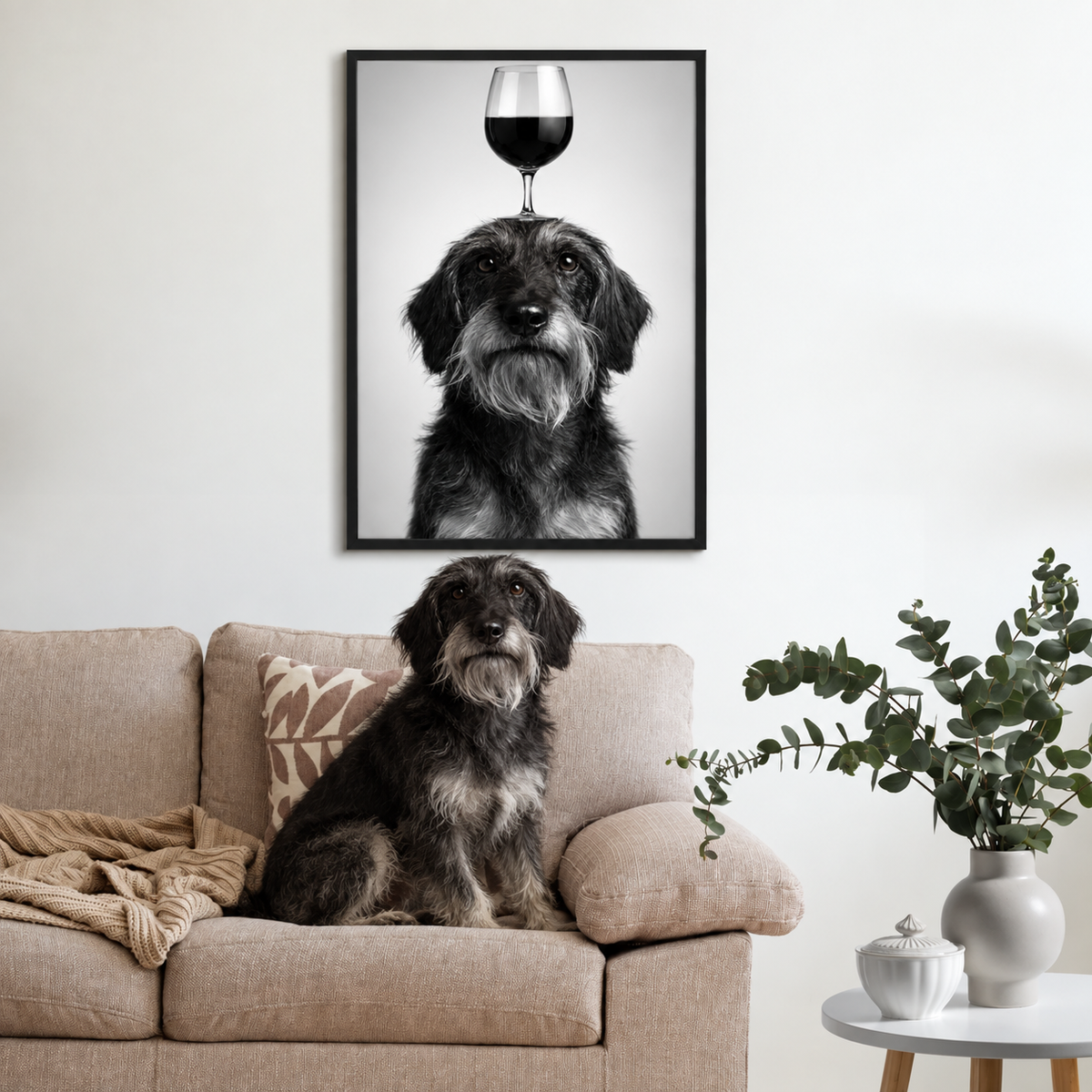 Black-and-white poster of a dog with a wine glass on its head, framed above a couch with cushions and plants.
