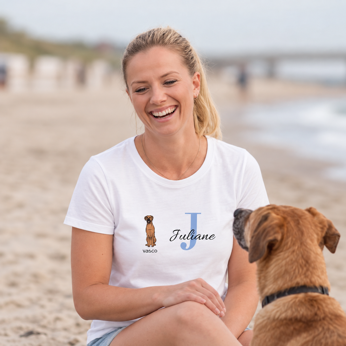 White T-shirt with a dog graphic and the name Juliane in blue letters. Woman sitting on the beach.