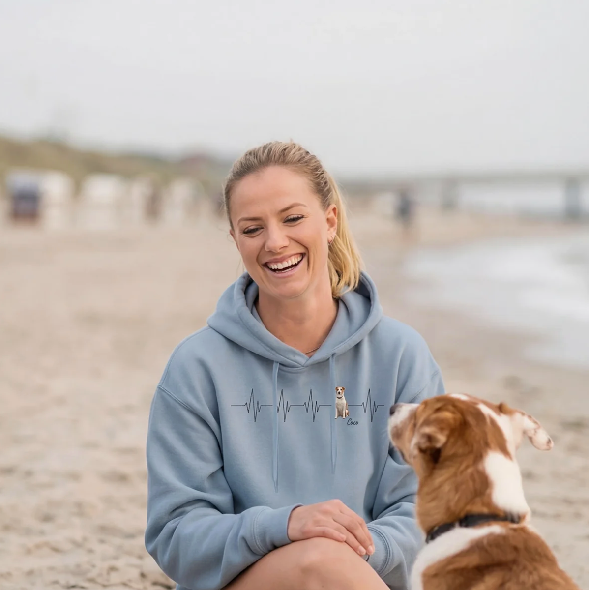 Personalized blue hoodie with heartbeat and dog motif, woman sitting on the beach smiling.