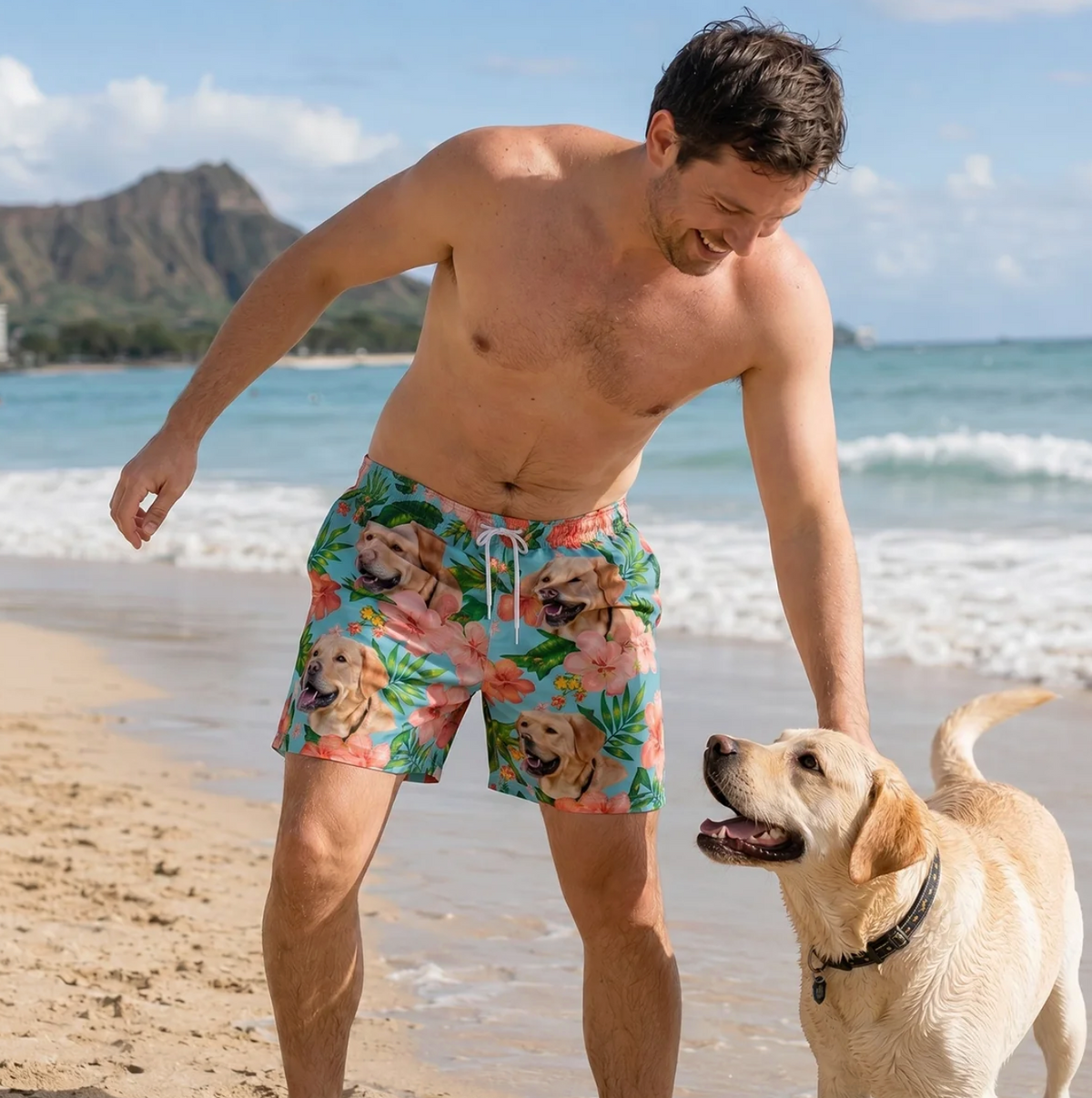 Man on the beach wearing colourful swim shorts with dog and flower design. Labrador playing in the sand next to him.