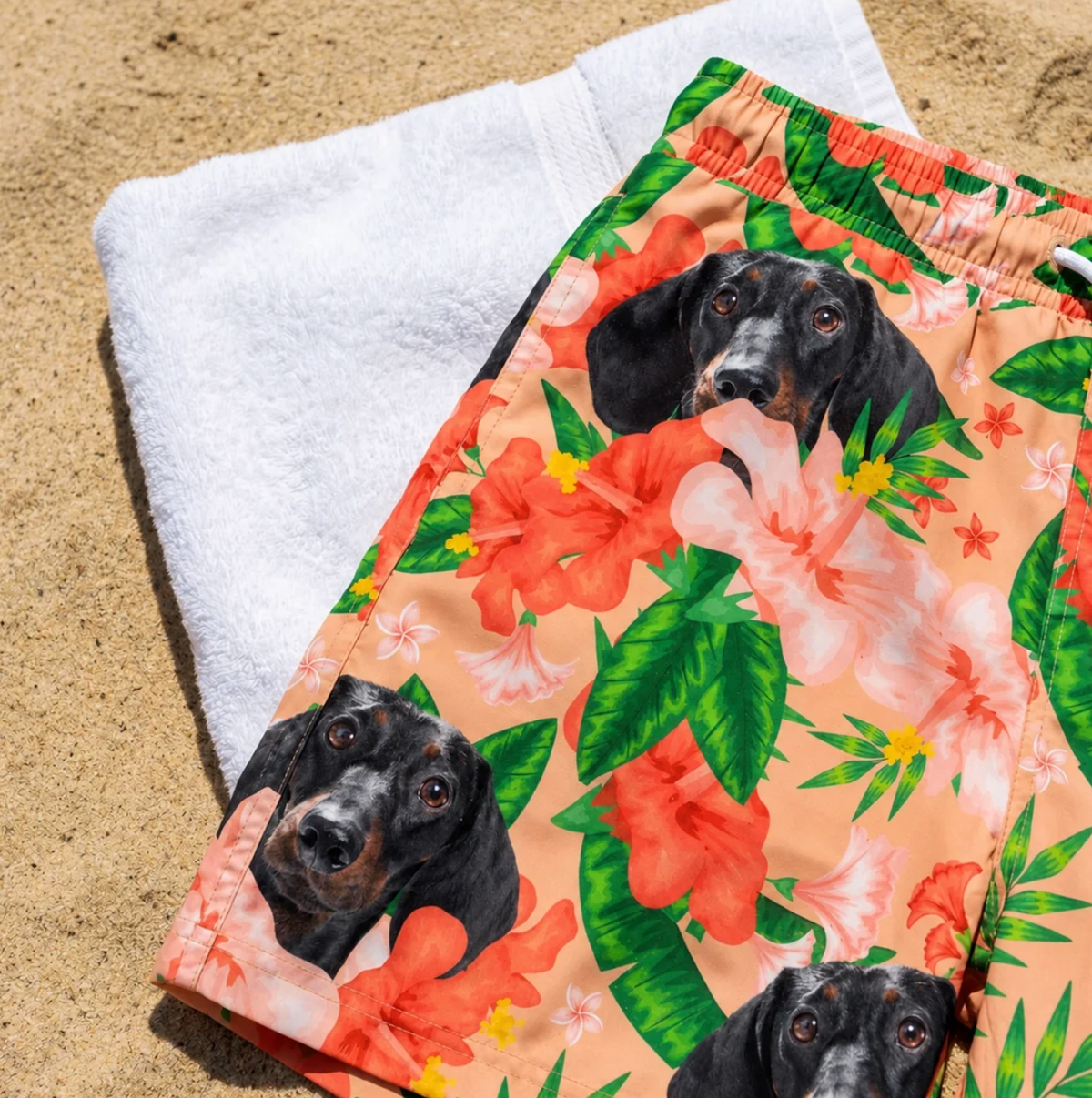 Personalized swim shorts with floral motifs and dog print lying on a sandy beach.