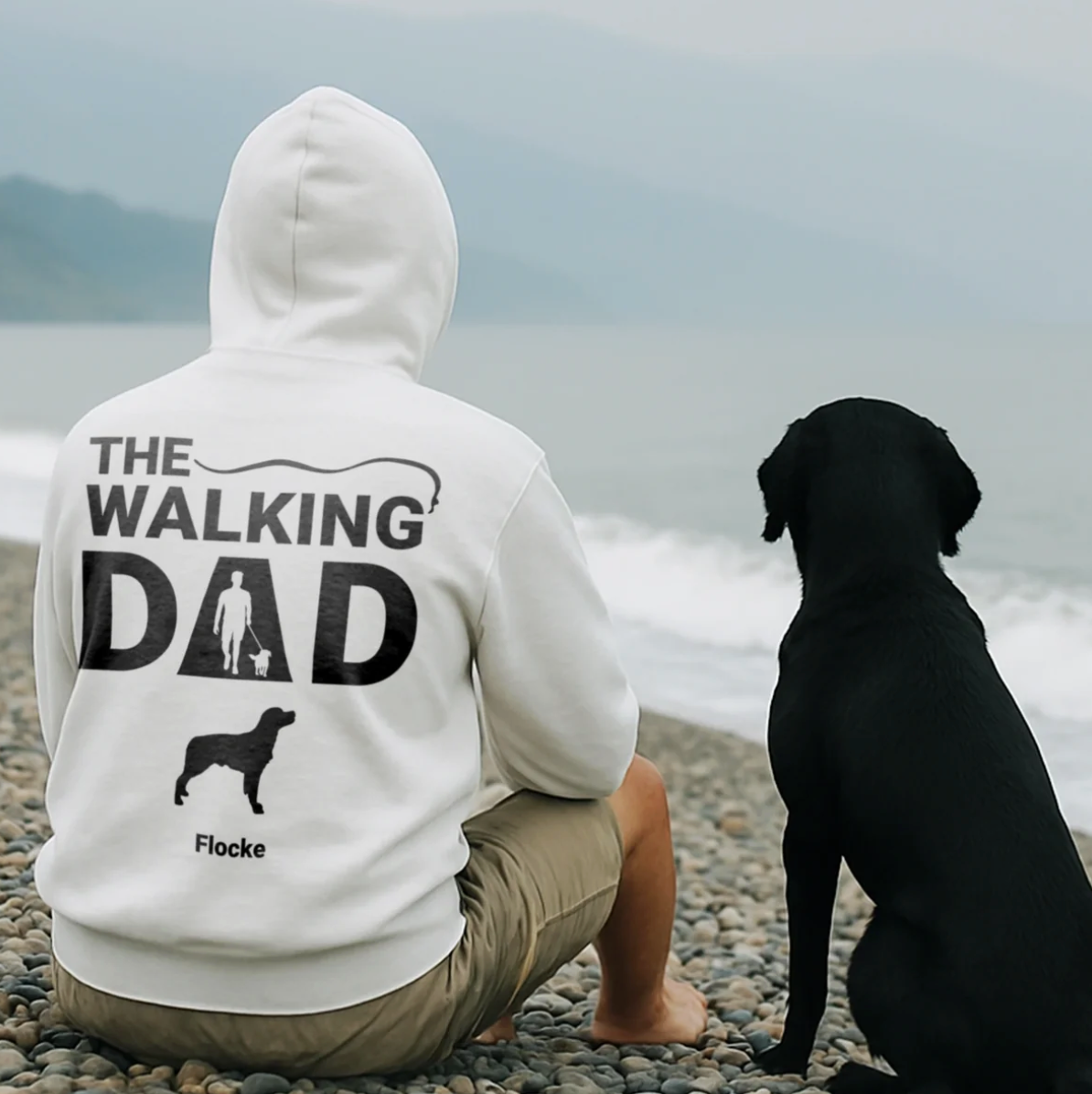 White hoodie with "THE WALKING DAD" back print and dog design, worn by a man sitting on the beach.